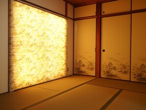A traditional Japanese room with tatami flooring, sliding doors with landscape paintings, and a window with floral blinds. - Lumiere De Bonnart (Okayama)