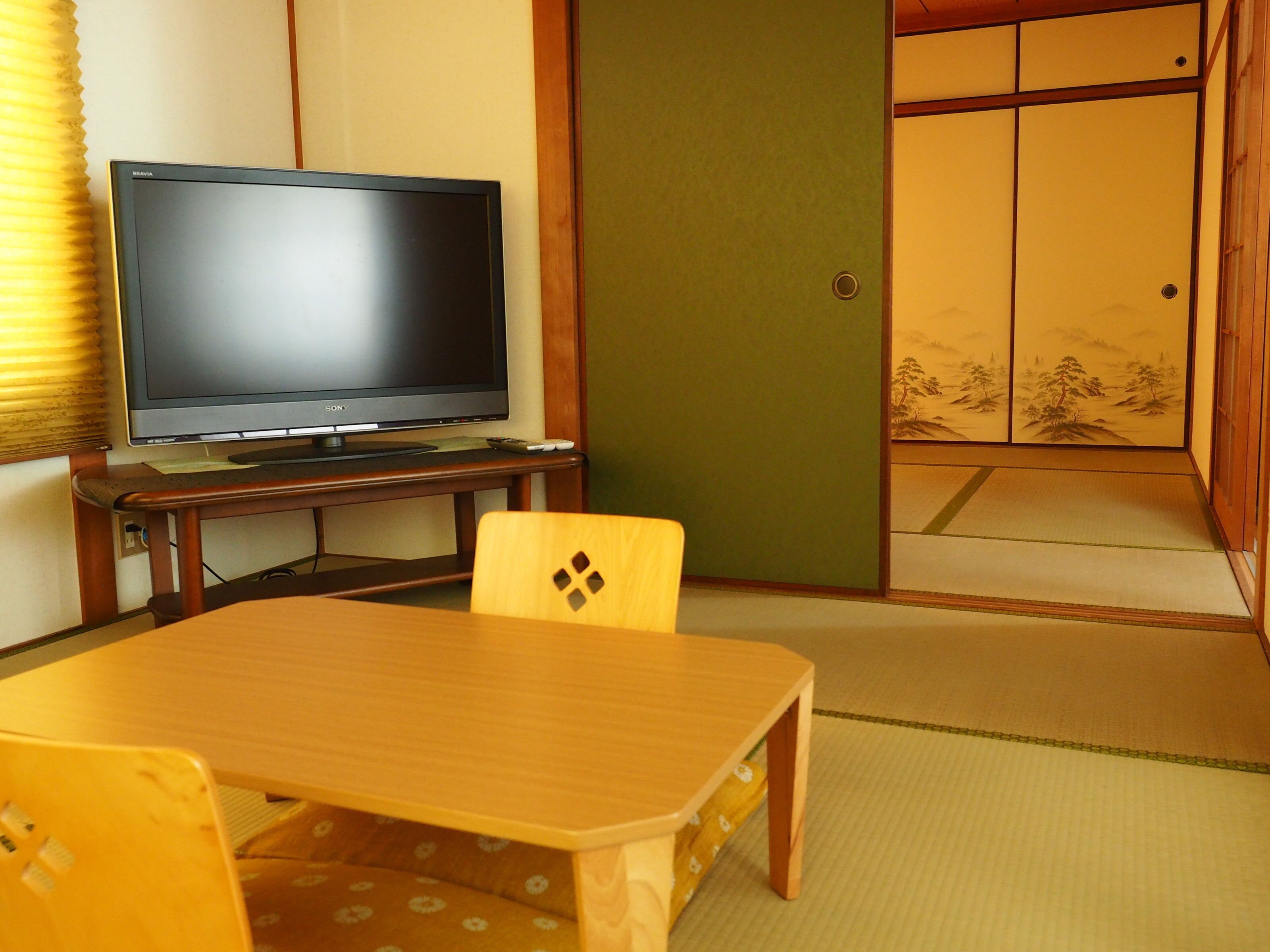 Um quarto japonês tradicional com tatames, uma mesa baixa, uma televisão sobre suporte e portas corrediças.