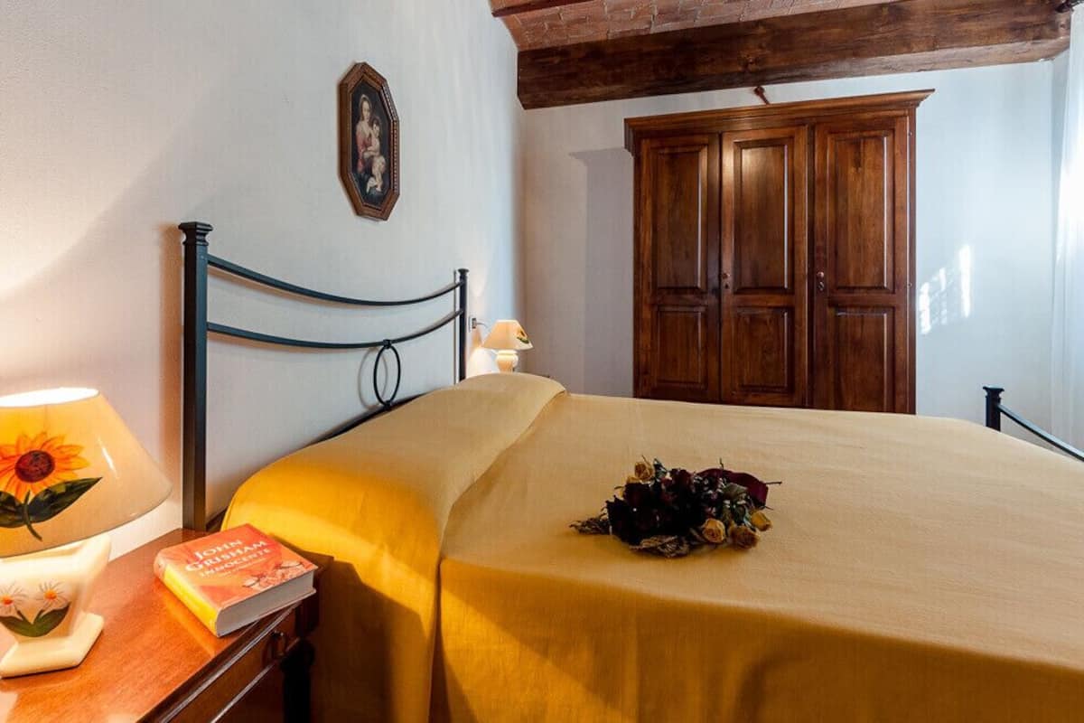 A neatly made bed with a yellow bedspread, a bedside lamp with a floral design, a framed picture on the wall, and a wooden wardrobe.