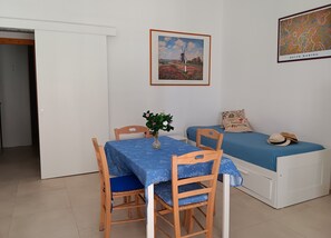 Une pièce comprenant une table à manger, des chaises, un lit avec une couverture bleue, un tableau accroché au mur et une porte blanche.