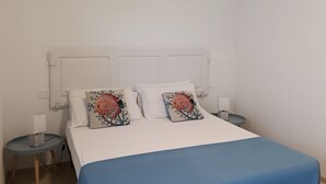 Una cama bien hecha con una colcha azul, dos almohadas decorativas y un cabecero blanco. Hay dos mesitas de noche con lámparas y una mesita auxiliar con una caja.