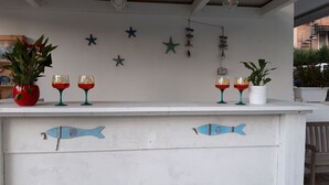 Eine Bar mit einer weißen Theke, verziert mit Sternfischen und einem Wandgemälde mit Fischen. Auf der Theke befinden sich zwei Kübelpflanzen und zwei Gläser Rotwein.