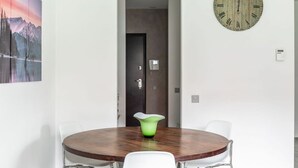Un espace de repas moderne avec une table ronde en bois, des chaises blanches et un vase vert servant de centre de table. Une grande horloge est accrochée au mur.