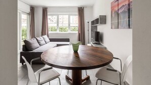 Un salon moderne avec une table ronde en bois, des chaises blanches, un canapé gris et une grande fenêtre avec des rideaux.