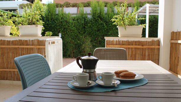 Una mesa dispuesta con dos tazas, una cafetera y un plato de pasteles en un entorno al aire libre con vallas de bambú y plantas en macetas.