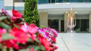 Een modern gebouw met een glazen ingang, een decoratieve metalen sculptuur en een rij kleurrijke bloemen.
