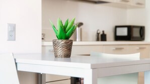 Een moderne keuken met een witte tafel, een geweven pot met een groene plant en een magnetron op de achtergrond.