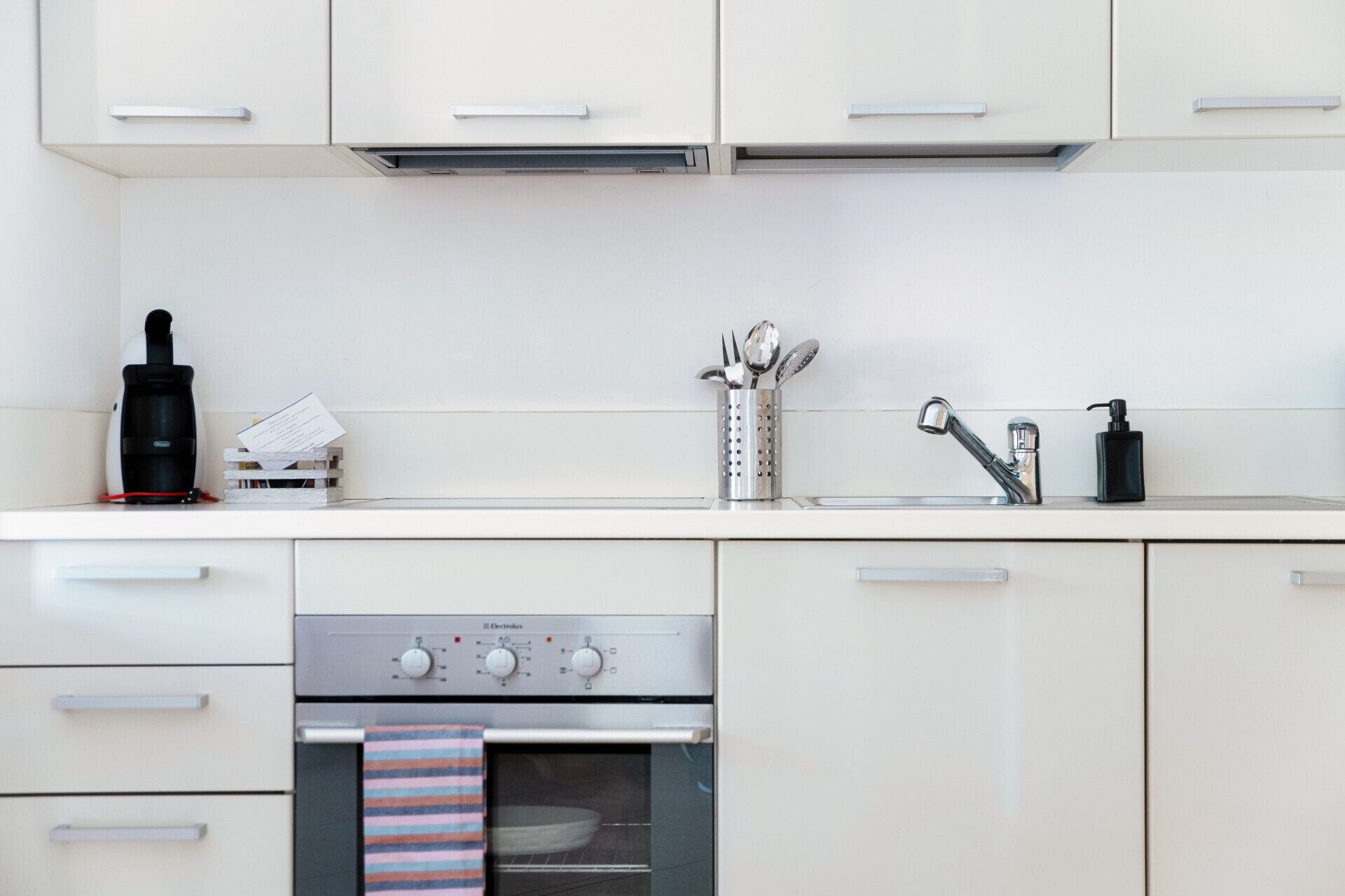 Una cucina moderna con mobili bianchi, una cappa in acciaio inossidabile, una macchina per il caffè, un bollitore, un set di utensili, un dispenser di sapone e un forno integrato.