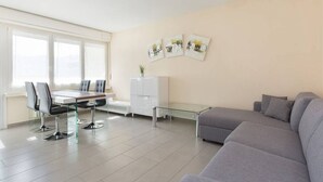 Una sala de estar moderna con un sofá gris, una mesa de vidrio para comer con cuatro sillas, un armario blanco y una planta pequeña.
