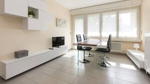 Una sala de estar moderna con una mesa de comedor, dos sillas, un televisor sobre un mueble blanco y una ventana con persianas.