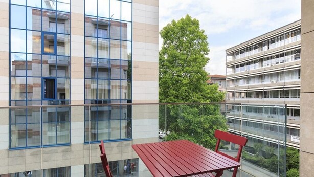 A balcony with a red table and chairs, overlooking a modern building with large windows and a tree in the background.