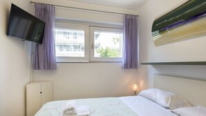 A hotel room with a bed, a flat-screen TV mounted on the wall, a window with curtains, and a bedside lamp.