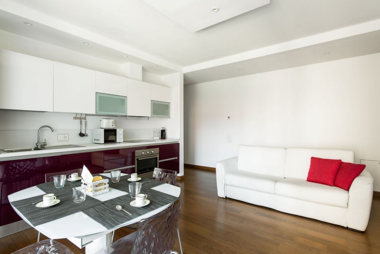 A modern kitchen with white cabinets, a dining table with chairs, and a white sofa with red cushions.