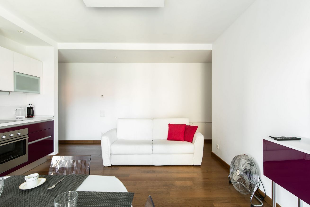 Een moderne woonruimte met een kitchenette, een witte bank met een rood kussen, en een eethoek met een tafel gedekt voor twee personen.