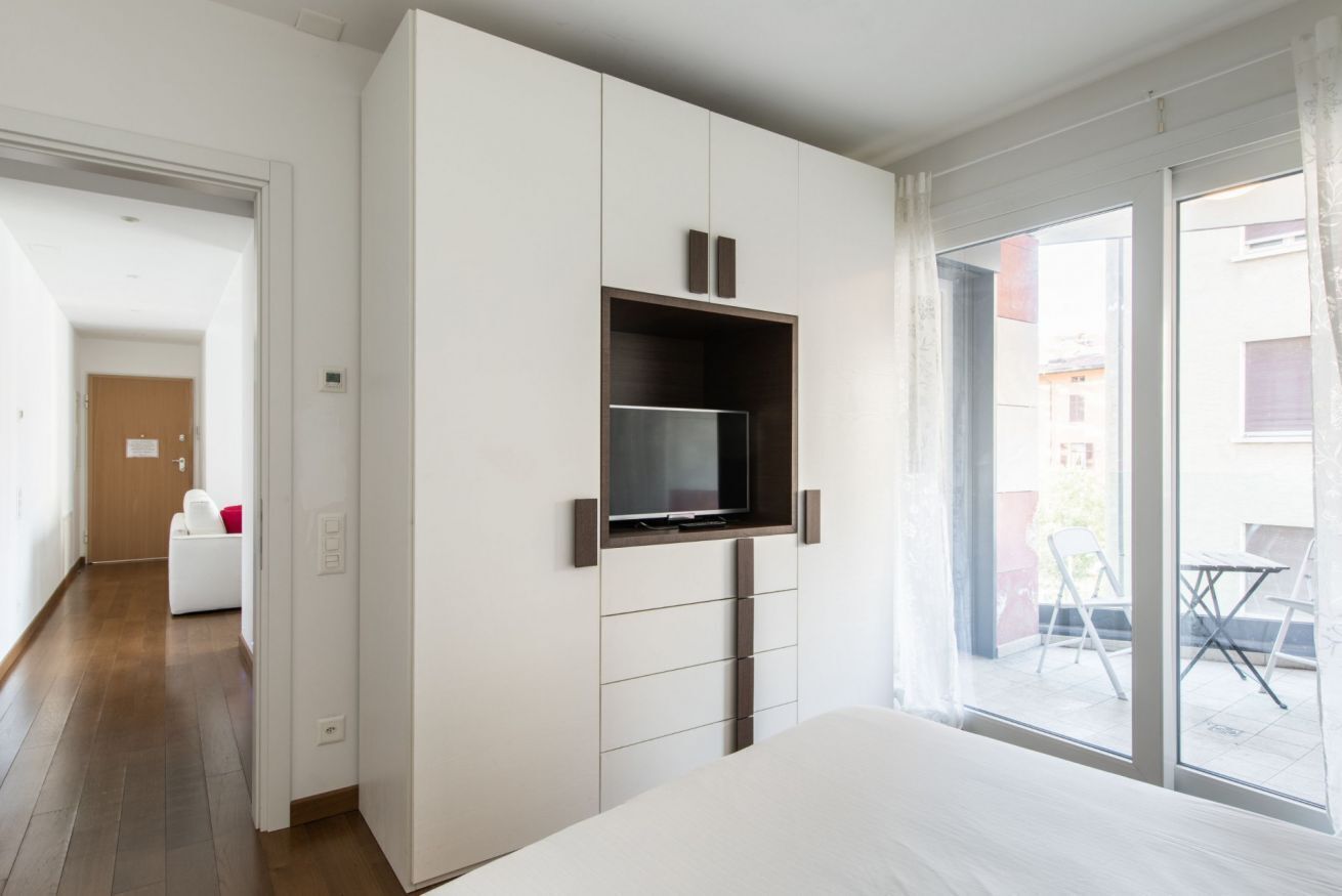 A modern bedroom with a large bed, a TV mounted on a white wardrobe, and a balcony with outdoor furniture.