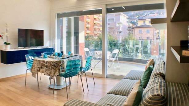 A modern living room with a sofa, a dining table with chairs, a TV, and a sliding glass door leading to a balcony.