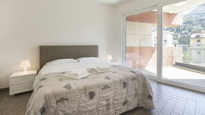 Ein Schlafzimmer mit einem Bett, Nachttischen und einem großen Fenster mit Blick auf Gebäude und Hügel.