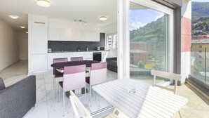 A modern kitchen with white cabinets, a dining area with a table and chairs, and a balcony with a view of buildings and hills. - Apartment two steps from the Exhibition Center of Lugano - ROGGIA 17 (Lugano)