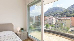 A bedroom with a large window, a bed, a bedside table with a lamp, and a view of a town and mountains. - Apartment two steps from the Exhibition Center of Lugano - ROGGIA 17 (Lugano)
