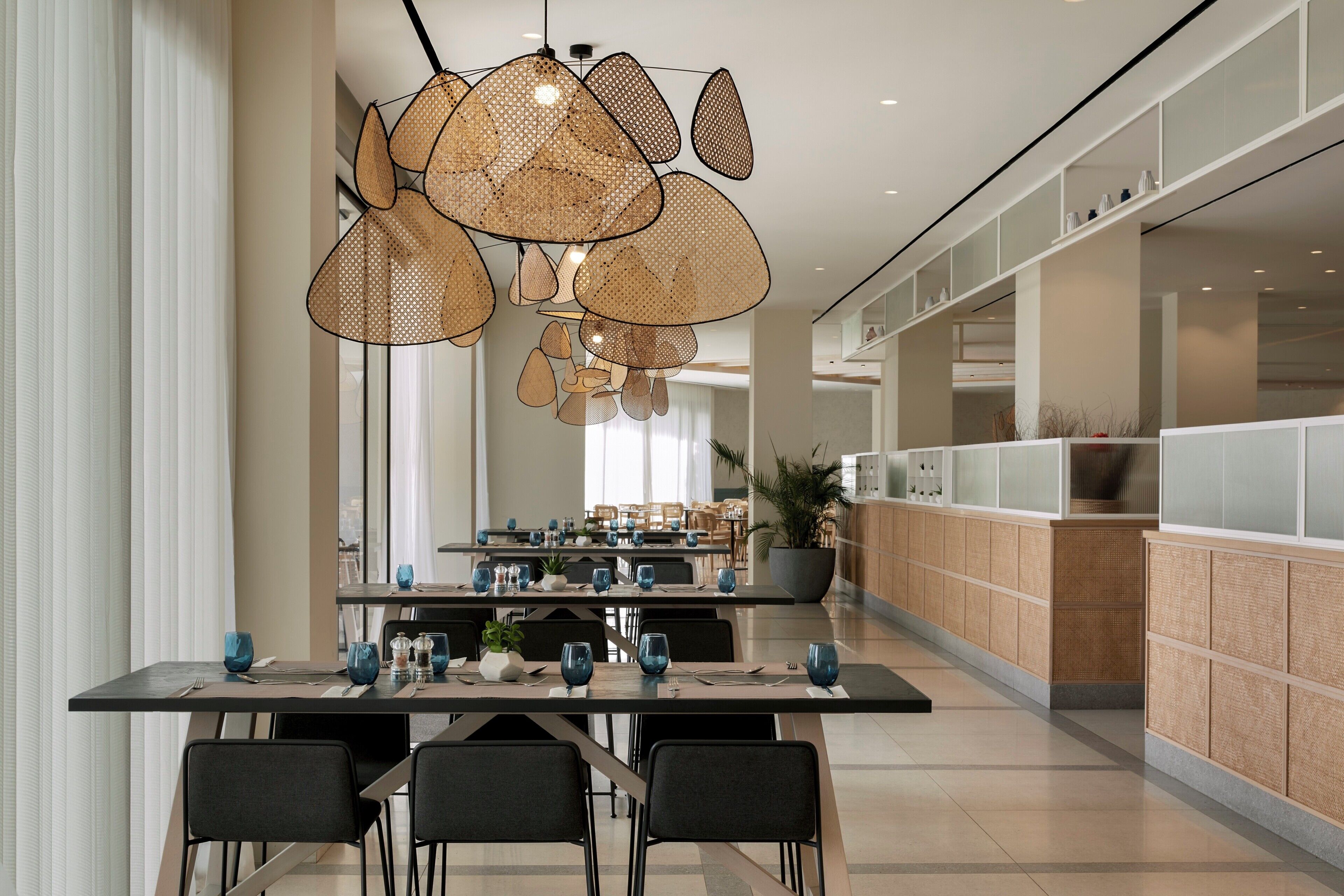 A modern dining area with a central hanging rattan chandelier, long tables, and chairs.