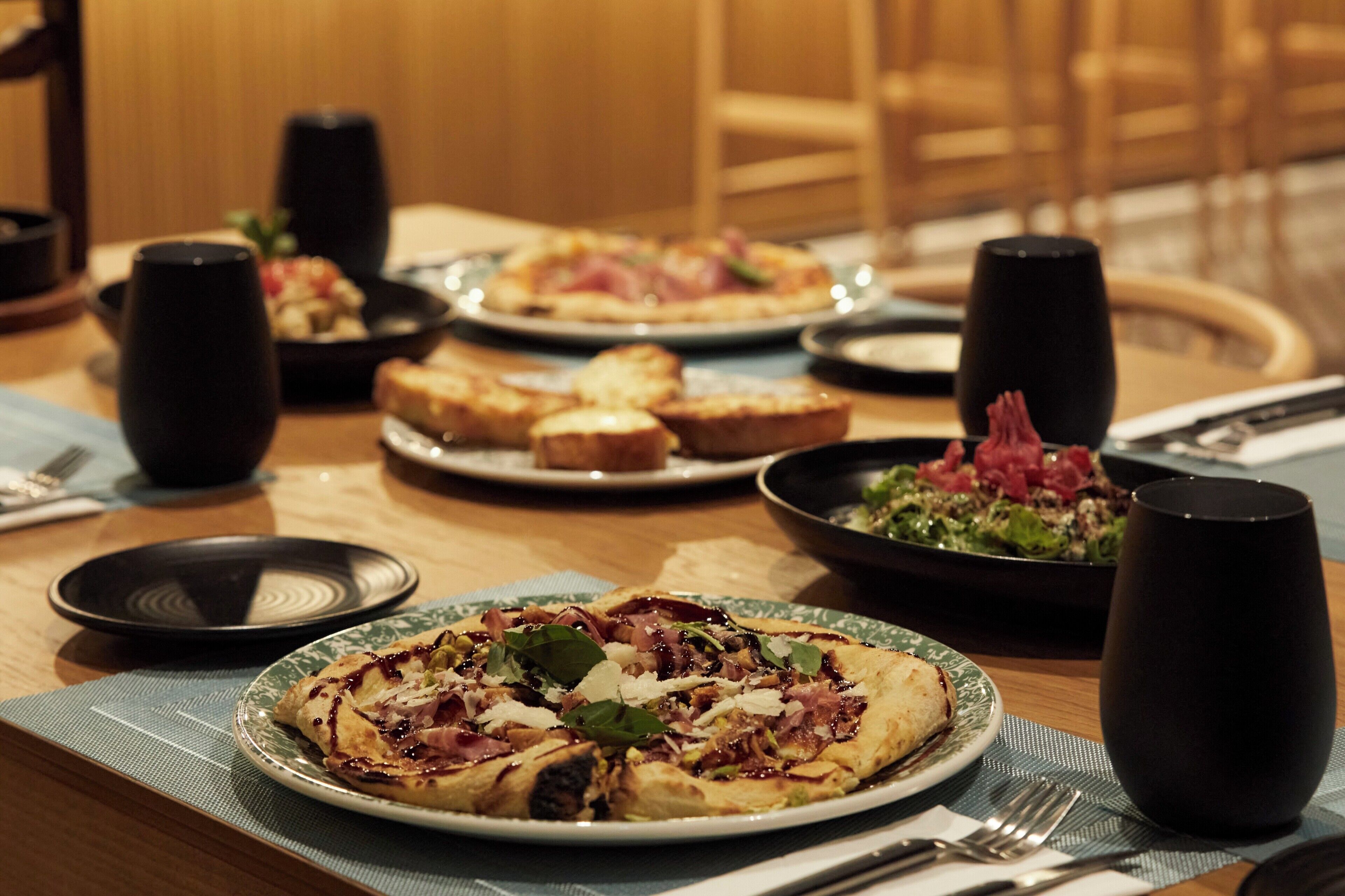 Ein Esstisch ist gedeckt mit Speisen, darunter Pizza, Brot und Salat, sowie schwarzen Tassen und Besteck.