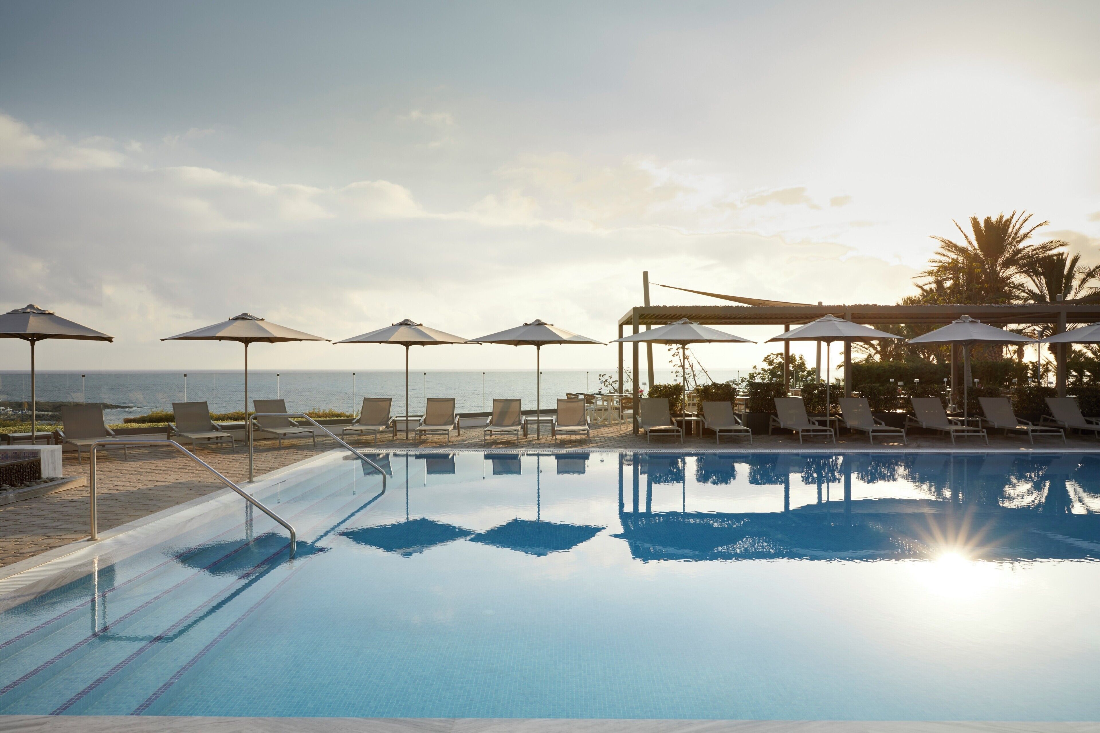 A swimming pool with lounge chairs and umbrellas, a clear sky, and a view of the sea.