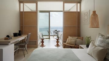 Quarto com uma cama grande, uma escrivaninha, uma cadeira, uma mesinha e vista para o mar através de uma porta deslizante.