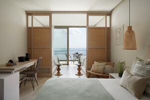 Un dormitorio con una cama grande, un escritorio, una silla, una mesita y vistas al mar a través de una puerta corredera.