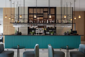 A bar area with a teal counter, multiple shelves of bottles, and hanging glasses. There are two small tables with chairs in front. - Mare Paphos (Kissonerga)