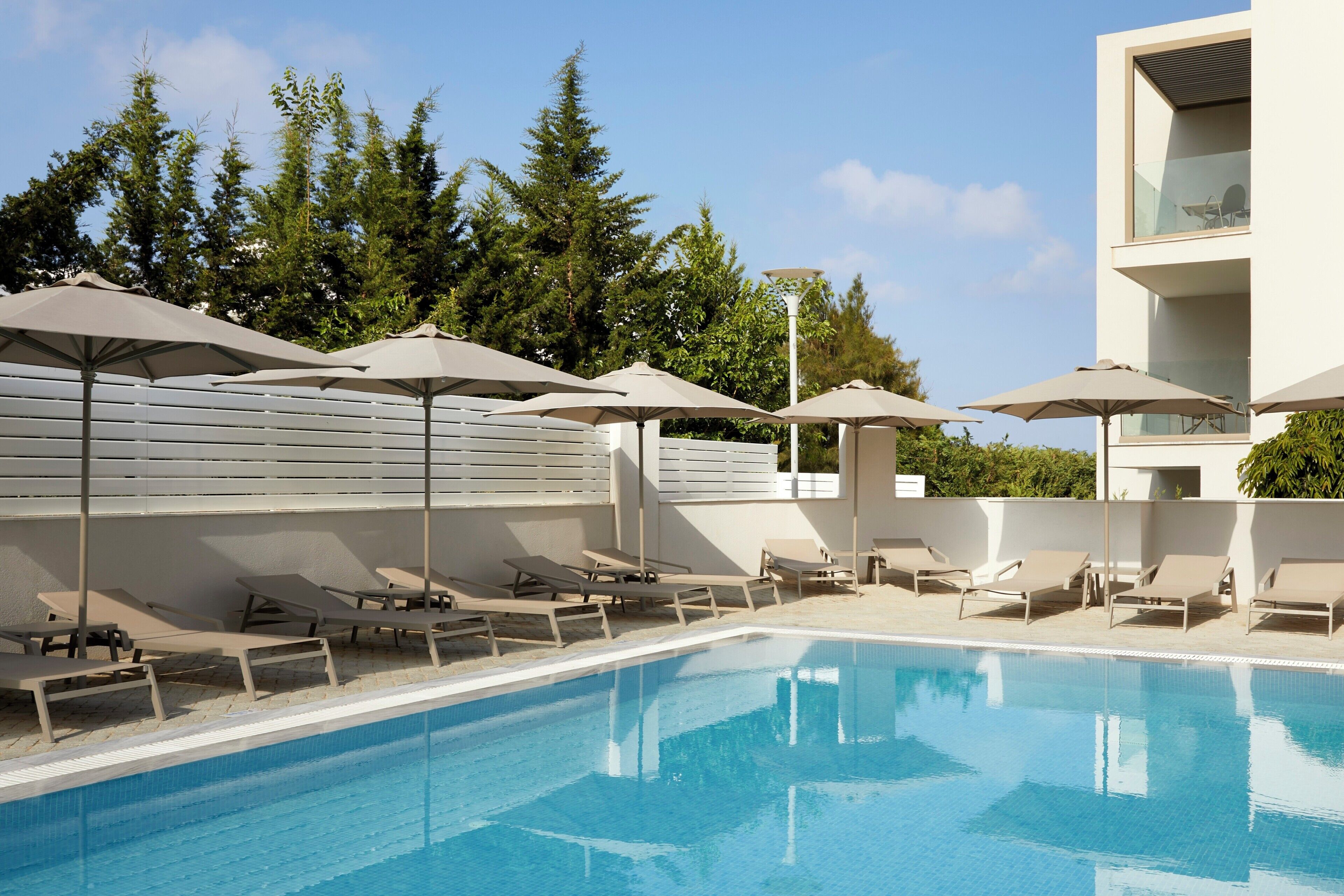 A swimming pool with lounge chairs and umbrellas, surrounded by trees and a modern building.