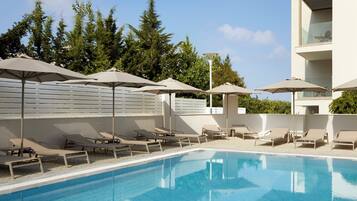 A swimming pool with lounge chairs and umbrellas, surrounded by trees and a modern building.
