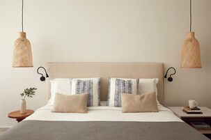 Una cama bien hecha con tablero cabecera color crema, dos lámparas de noche y una mesita de noche con un libro y una taza.
