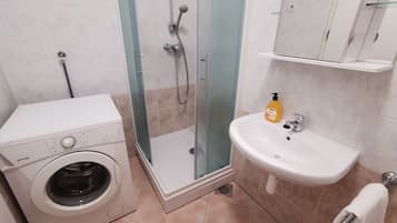 A compact bathroom with a washing machine, a glass-enclosed shower, and a sink with a bottle of soap.
