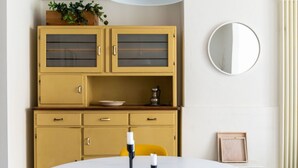 Une pièce avec un miroir rond, un meuble jaune aux portes vitrées, une table blanche avec des bougies et une chaise jaune.