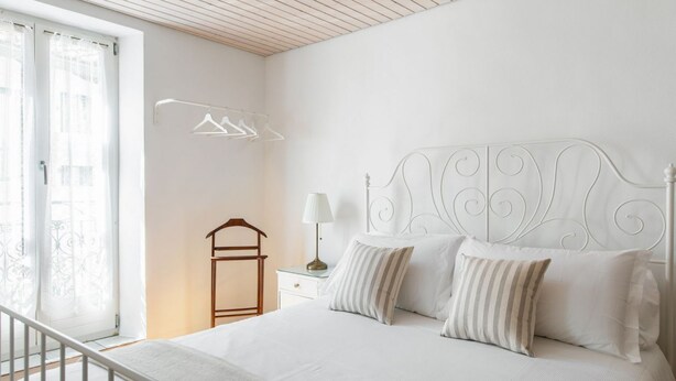 Une chambre à coucher avec un lit blanc, une table de chevet avec une lampe, une chaise en bois et un portant à vêtements.