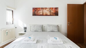 Hotellihuone, jossa on sänky, metsämaisemaa esittävä taulu, patteri, yöpöytä lampun kanssa ja puuovi.
