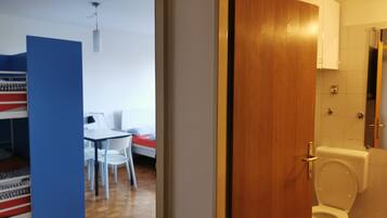 Une pièce avec un lit superposé, une petite table avec des chaises et une porte en bois donnant sur une salle de bain équipée de toilettes et d’une douche.