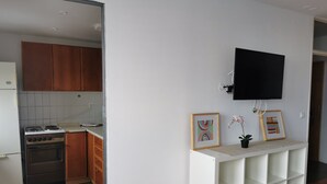 A compact kitchen with wooden cabinets, a stove, and a white countertop. A white shelving unit with a flat-screen TV above it is visible. - Dunajska Apartment (Ljubljana)