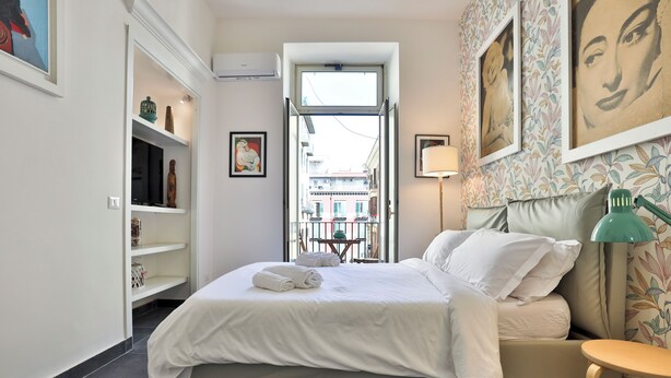 Une chambre à coucher avec un lit, une table de chevet, une lampe, une fenêtre et une vue sur la rue.
