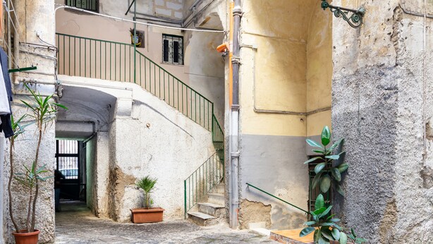 Une ruelle étroite avec un escalier menant à l’étage supérieur, des plantes en pot et un lampadaire.