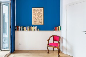 Ein Zimmer mit blauer Wand, einem hölzernen Stuhl mit rotem Kissen, einem Bücherregal voller Bücher und einem gerahmten Bild.