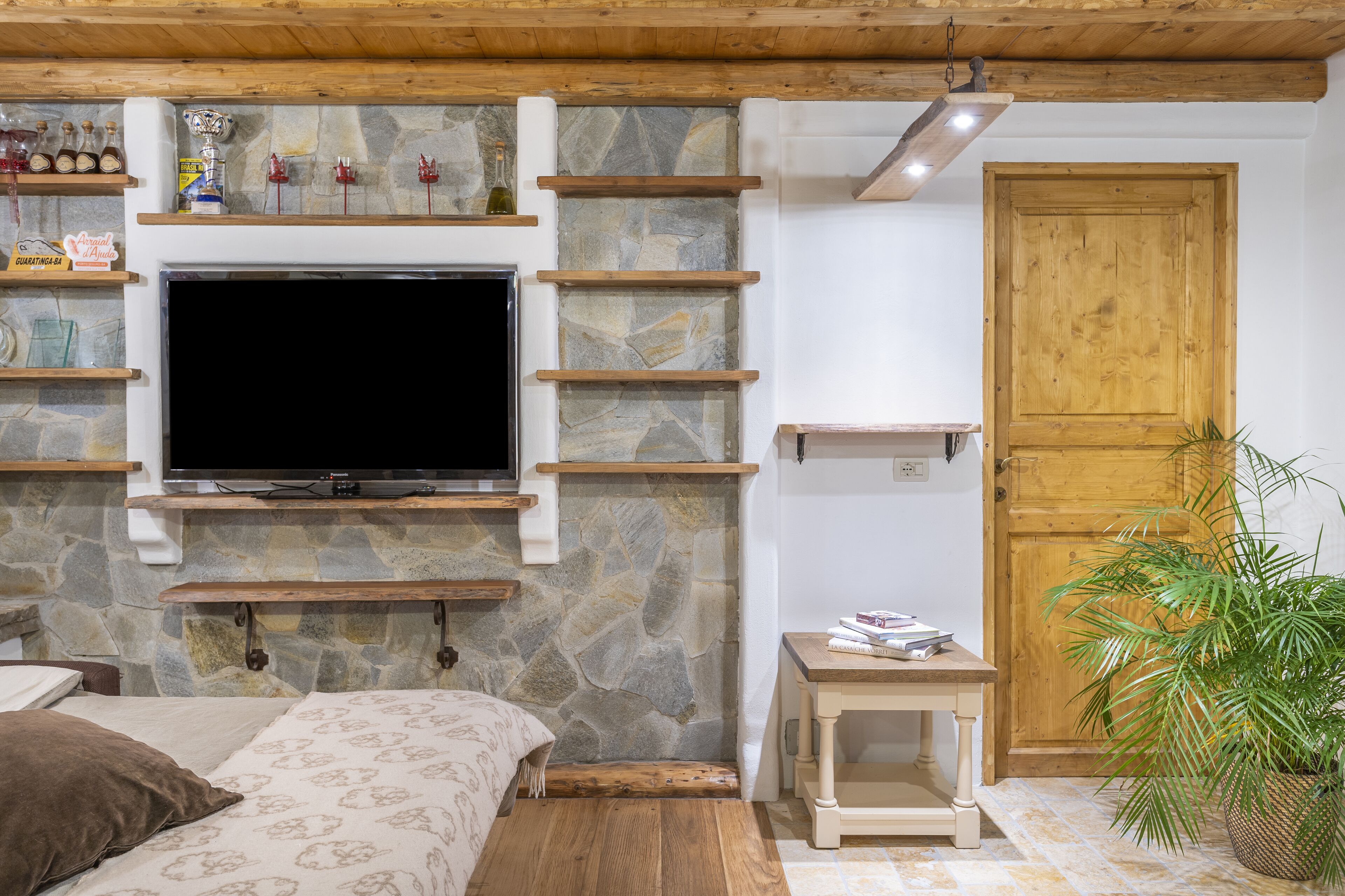 Ein gemütlicher Raum mit Kamin aus Stein, Holzregalen, einem Fernseher, einem Bett, einem kleinen Tisch und einer Tür.