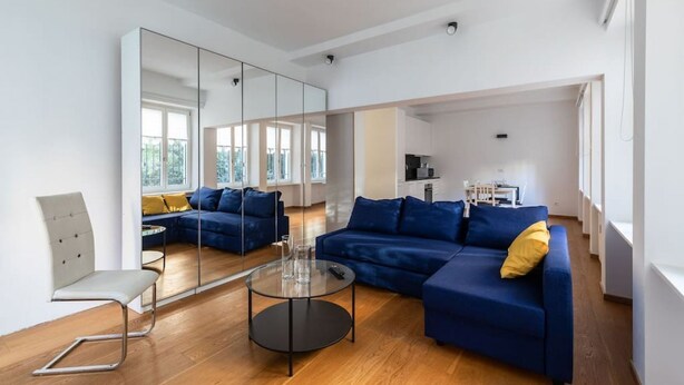Un salon moderne avec un canapé bleu, une table basse en verre et un mur miroitant qui reflète la pièce.