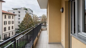 En balkon med fliset gulv, sort gelænder og udsigt til omkringliggende bygninger.