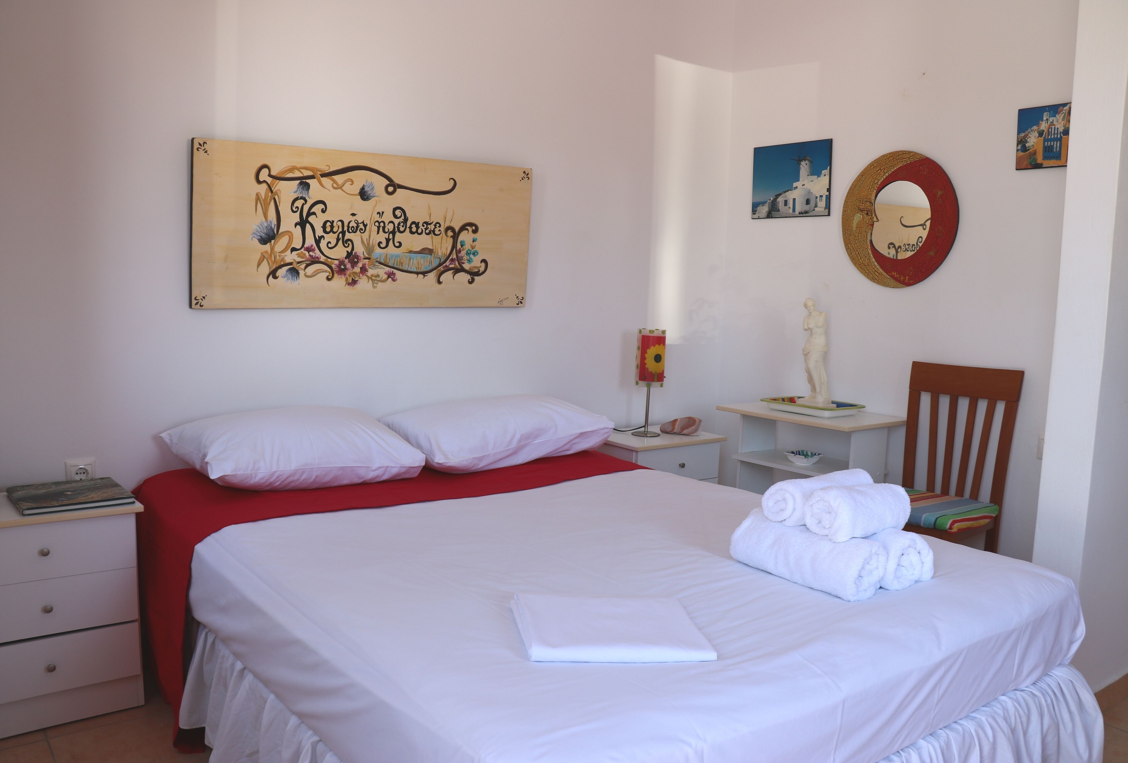Una cama bien hecha con sábanas blancas, un plaid rojo y una toalla doblada. Una mesita de noche con una lámpara y un adorno. Una silla de madera está colocada junto a la cama.