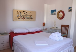 Una cama bien hecha con sábanas blancas, un plaid rojo y una toalla doblada. Una mesita de noche con una lámpara y un adorno. Una silla de madera está colocada junto a la cama.