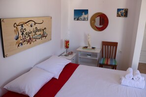 A neatly made bed with white and red bedding, a small bedside table with a lamp, a chair with a colorful cushion, and a wall with a decorative mirror and framed pictures. - Flora's Red Studios (Milos)