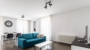 A modern living room with a blue sofa, a white coffee table, a dining area with chairs, a clock on the wall, and a radiator.