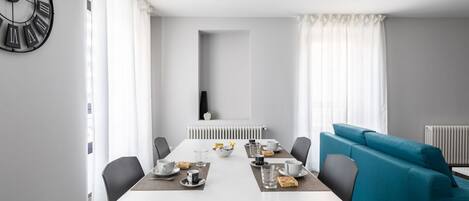 Un coin repas moderne avec une table blanche dressée pour deux personnes, un canapé bleu et une horloge murale noire.