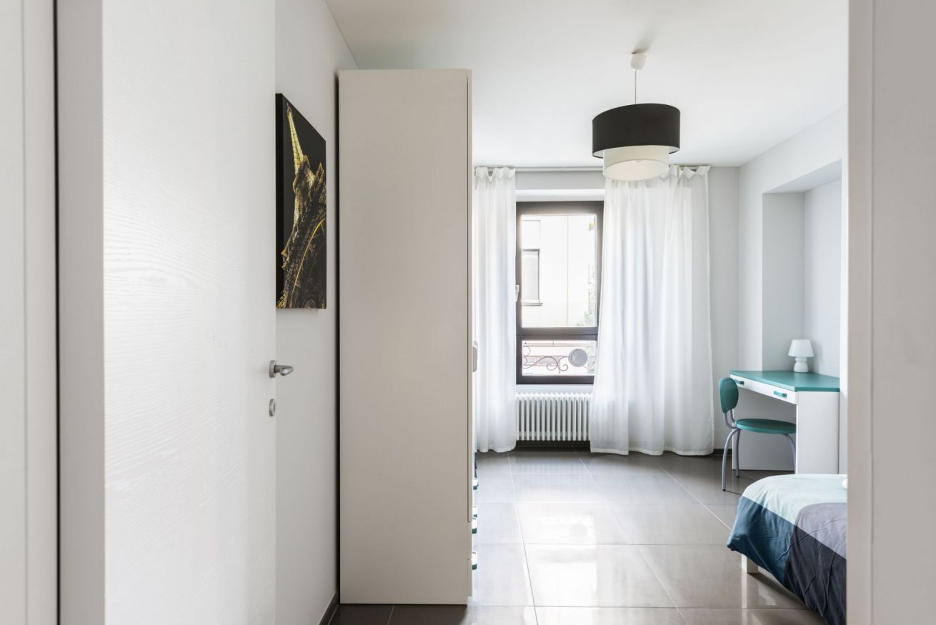 Habitación de hotel con cama, escritorio y silla. Se observa una ventana con cortinas y un radiador.